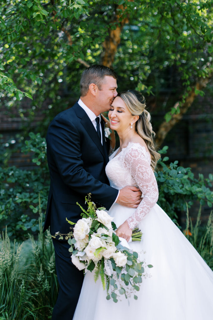 Groom kisses his bride’s head with a loving smile while embracing her, capturing a heartfelt moment on their wedding day. Toly Dzyuba Photography.
