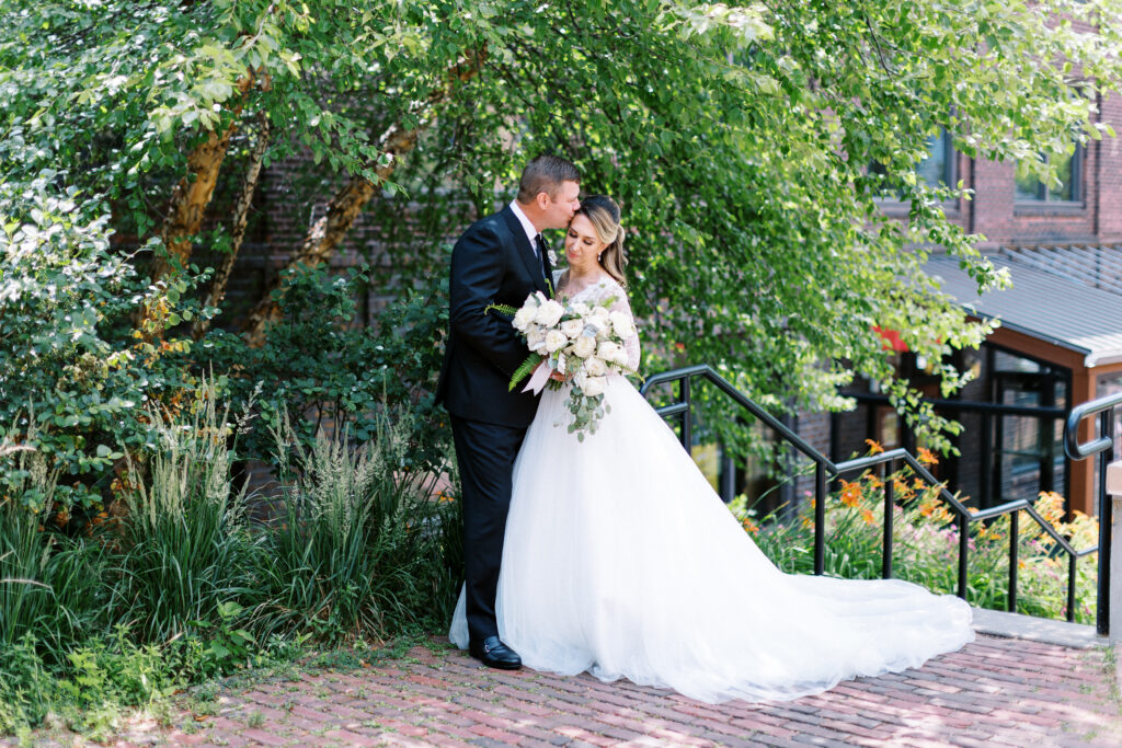 Bride holding flowers in a white dress with groom giving her kiss on the head.