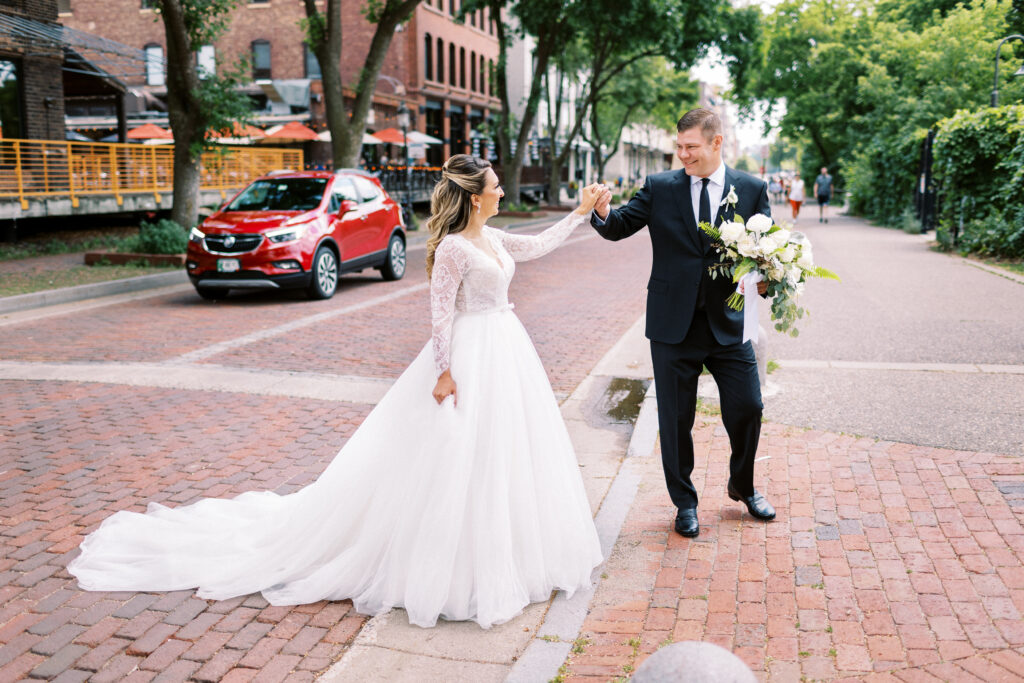 Bride and groom cross the street in Minneapolis. Bride and groom are walking across the street hand-in-hand. The groom has his hand in her hand and guiding her.