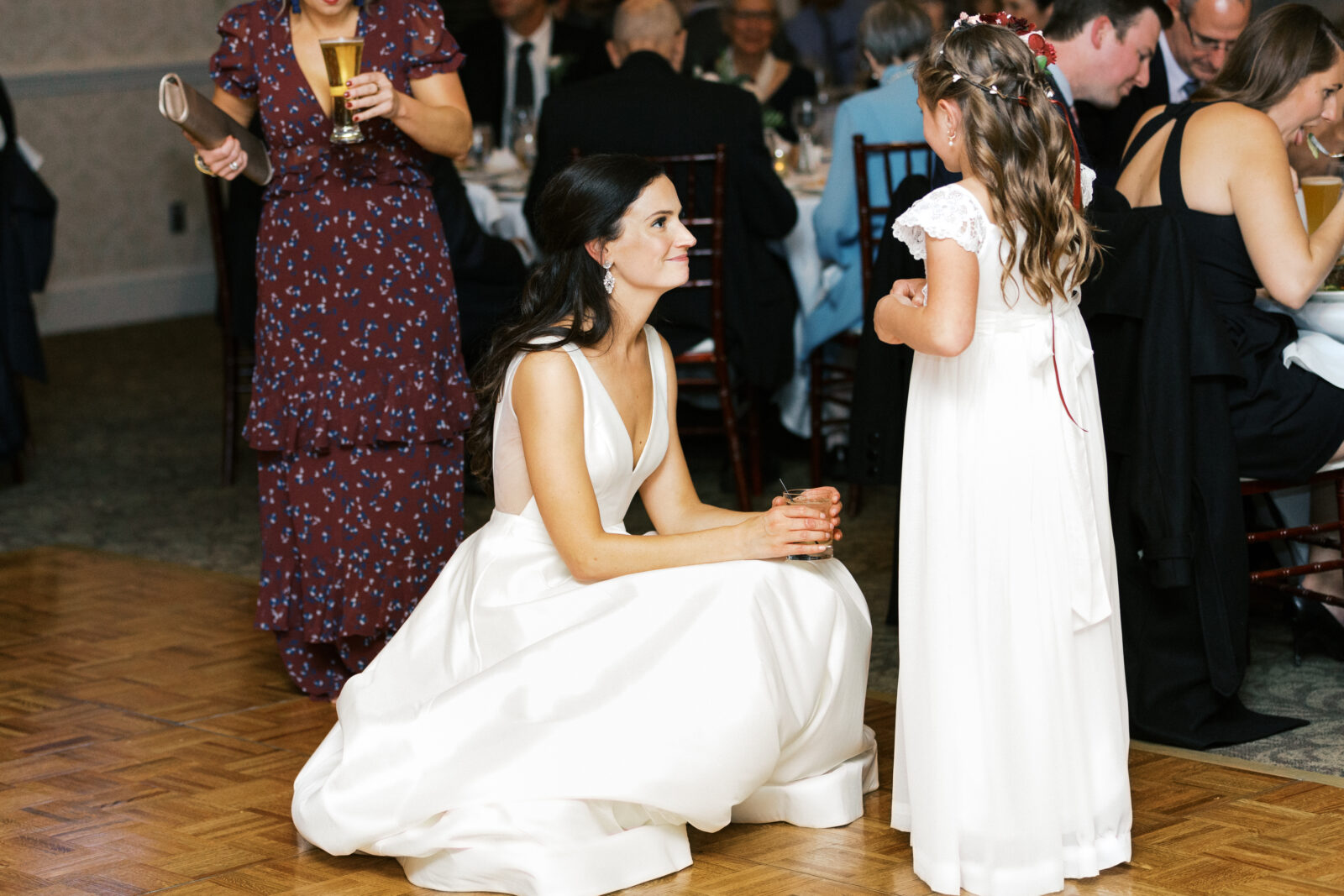 The bride is talking to her flower girl. The bride is wearing a white dress. The flower girl is wearing a white dress and a flower crown.