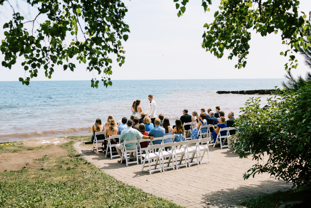 Toly Dzyuba Photography captures a moment during a Minnesota North Shore wedding ceremony. Guests, in casual wear, are gathered in a small group on the beach near the edge of Lake Superior. The bride and groom stand at the front of the crowd, with the groom in a suit and the bride in a long white gown. A serene, summer day is portrayed, as the sky is clear and the lake water is a calm blue.