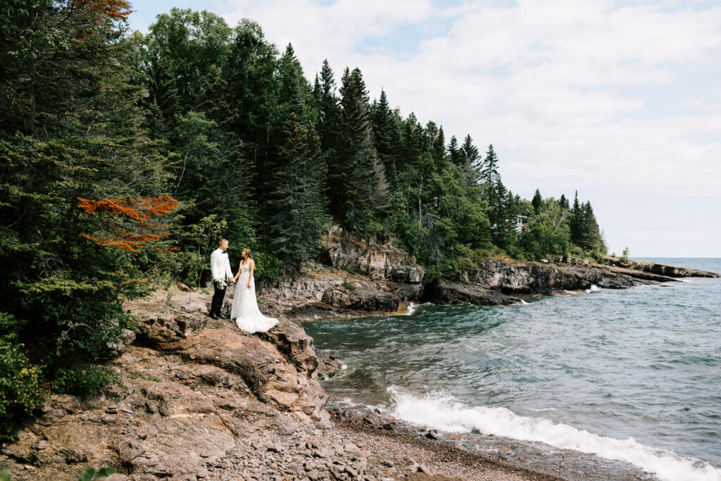 Bride and groom holding hands and looking at each other with a forest and Lake Superior in the background, captured by Toly Dzyuba Photography.