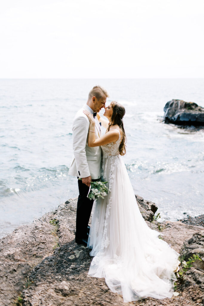 Newlyweds standing together on a rock along the North Shore, captured by a Minnesota wedding photographer, with the scenic lake and shoreline in the background.