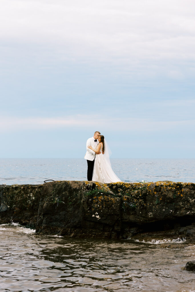 Groom is kissing the bride and embracing while standing on a rock along the North Shore of Lake Superior.