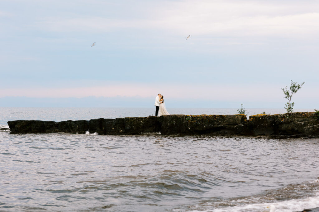 Bride and groom standing on rocky shoreline, sharing a kiss with a peaceful lake superior in the background.