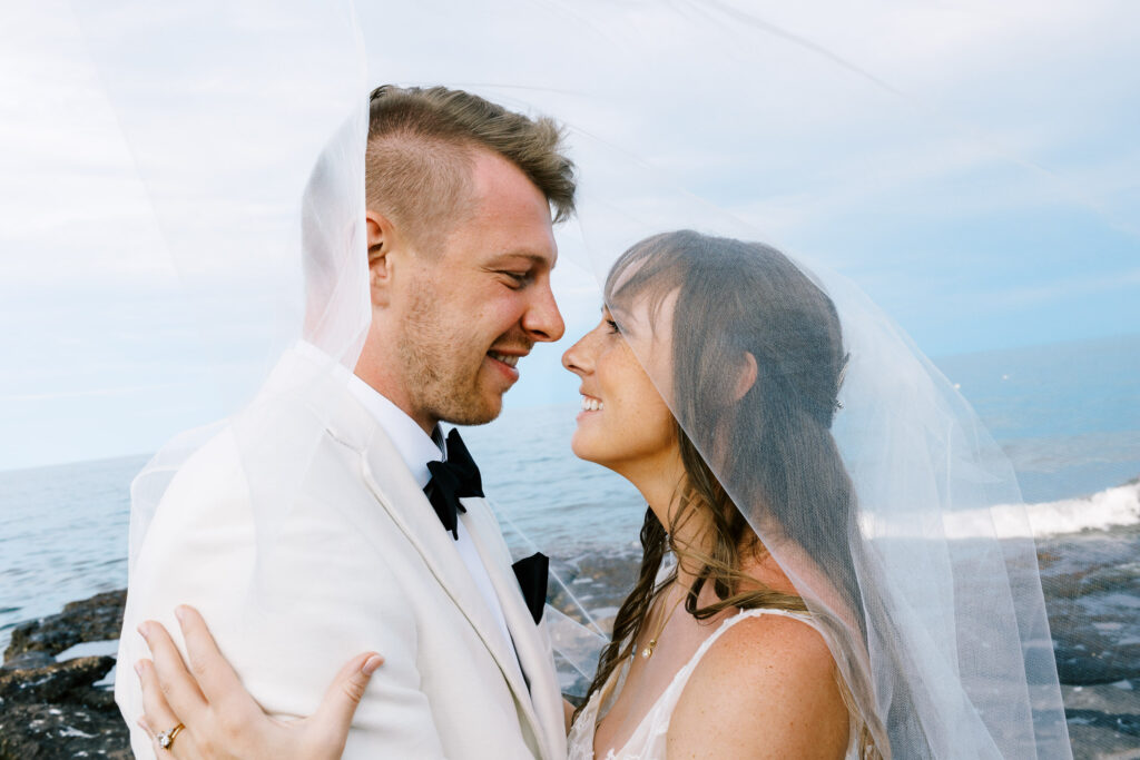 A close-up shot by Toly Dzyuba Photography of a bride and groom on the North Shore, smiling warmly as they gaze into each other's eyes, with Lake Superior in the background.