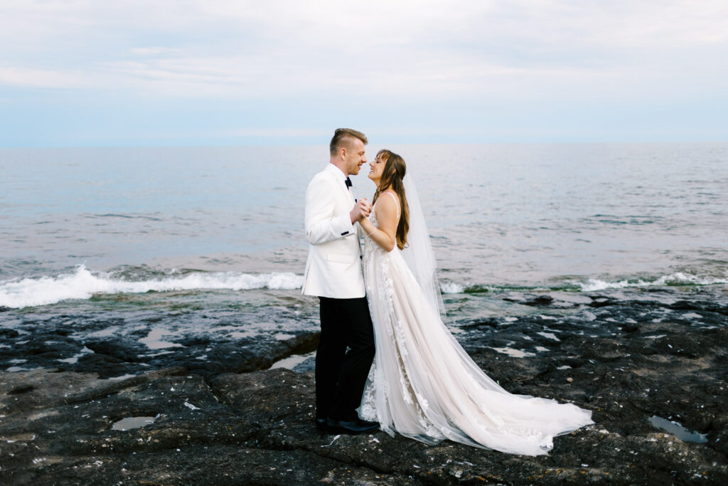 A close-up shot by Toly Dzyuba Photography of a bride and groom on the North Shore, smiling warmly as they gaze into each other's eyes, with Lake Superior in the background.