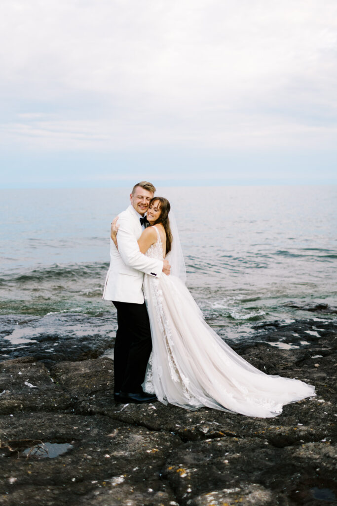 Bride and groom embracing with closed eyes on the North Shore of Lake Superior, with the serene lake and rocky shoreline in the background.