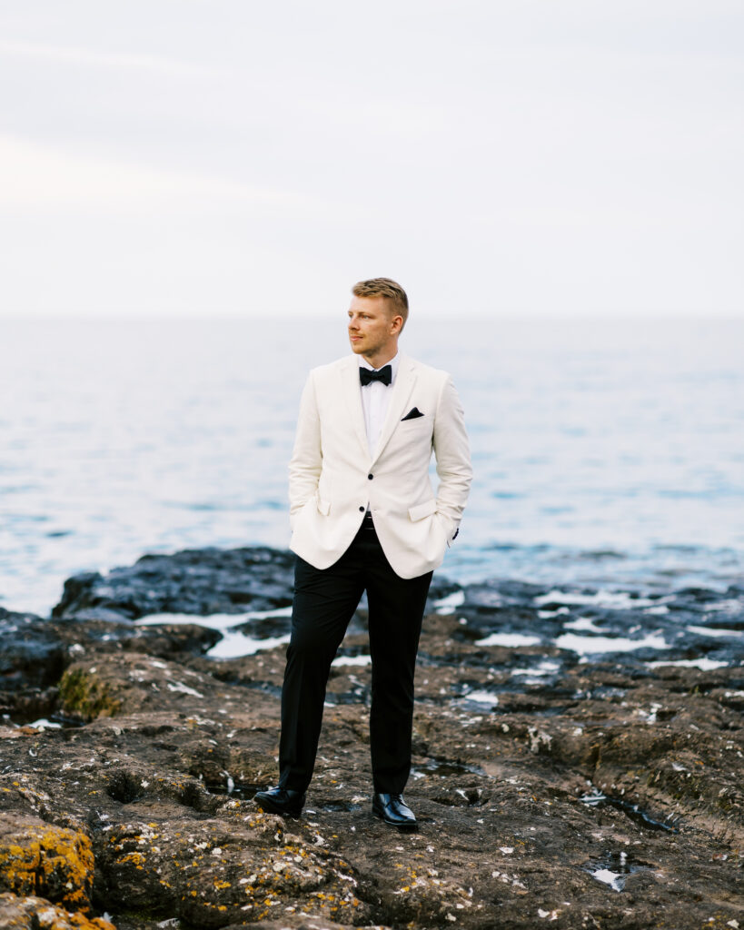 Groom poses with his hands in his pocket and looks away at the Lake Superior.