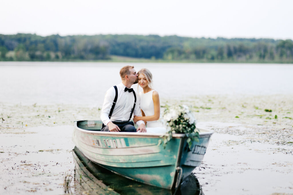 Groom on the lake is kissing his bride in the chick while in the boat.