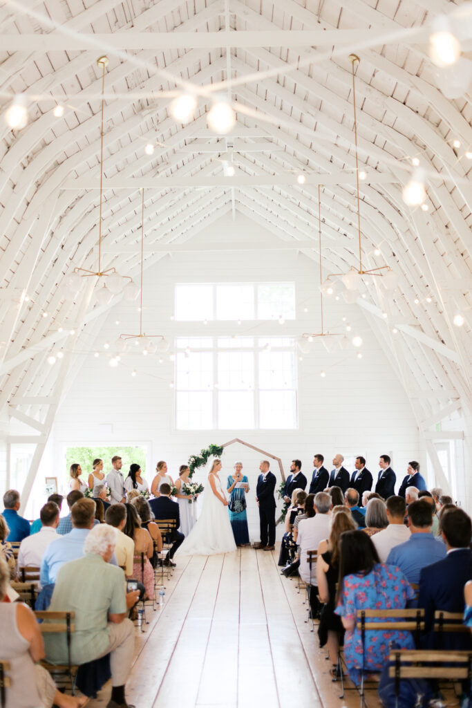 A wide shot of a wedding ceremony in the white barn at Ivory North. The bride and groom are standing at the altar, and the guests are seated in the pews.