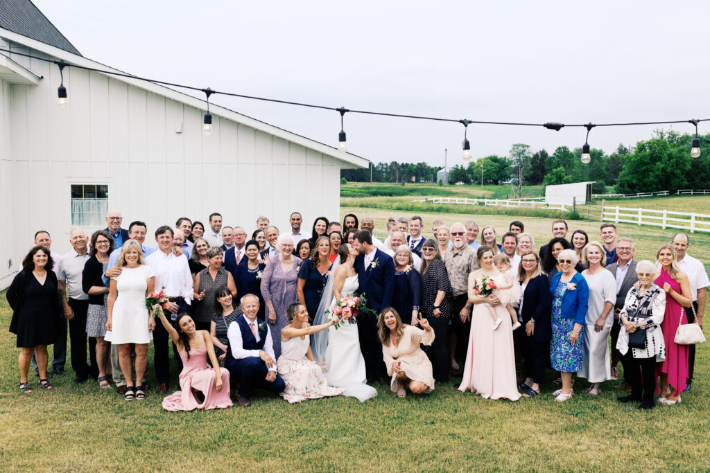 Wedding guests celebrating with a loud joy during a large group photo as the bride and groom share a kiss at Abella Weddings and Events in Minnesota.