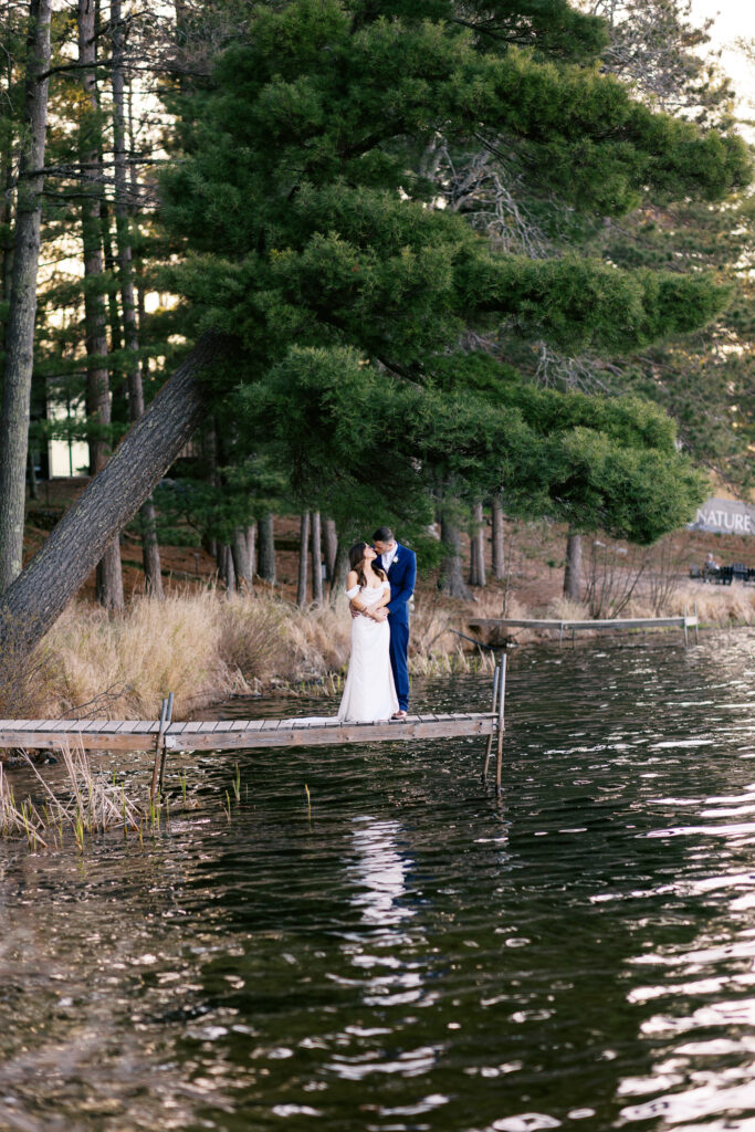 The Bride and Groom standing on a dock, hugging and kissing, with green pine trees in the background.