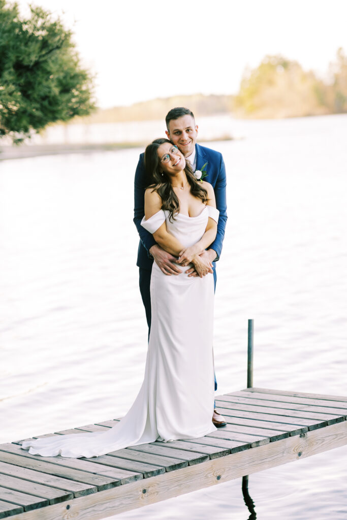 In the stillness of a gorgeous day, newlyweds shared a tender embrace as they looked out over a placid lake. This moment was made permanent in a photograph by Toly Dzyuba Photography.