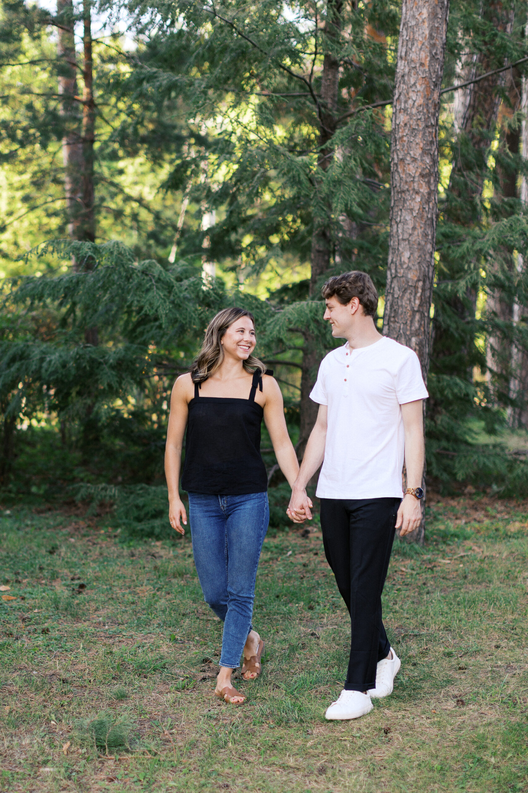 The couple walks through the woods, holding hands and smiling as they look at each other, radiating love. Captured by Toly Dzyuba Photography, your trusted Minnesota wedding photographer.