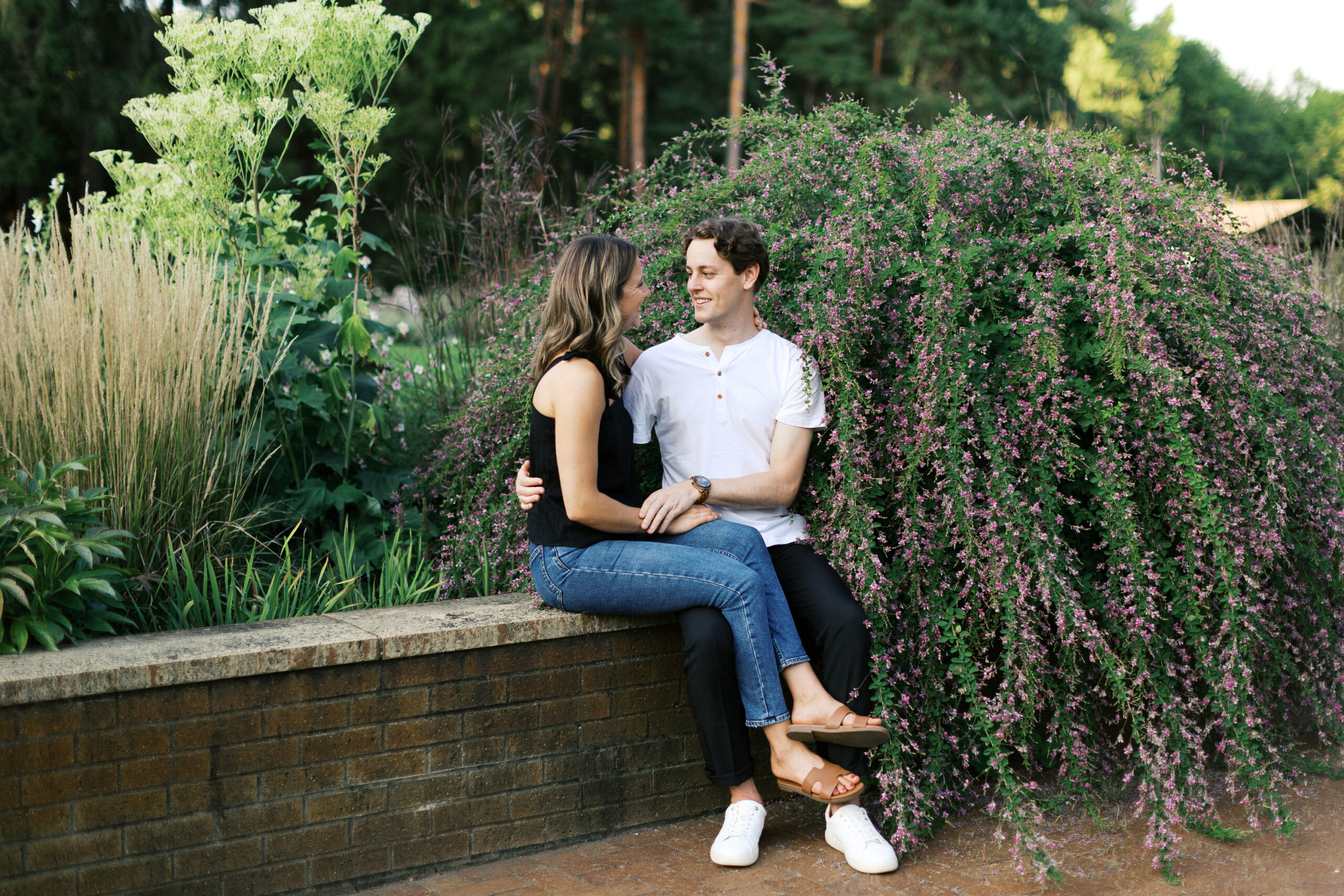 Sitting at Noerenberg Gardens on the edge of a garden wall, the couple looks at each other with a smile, sharing intimate moment. Captured by Toly Dzyuba Photography, your trusted Minnesota wedding photographer.