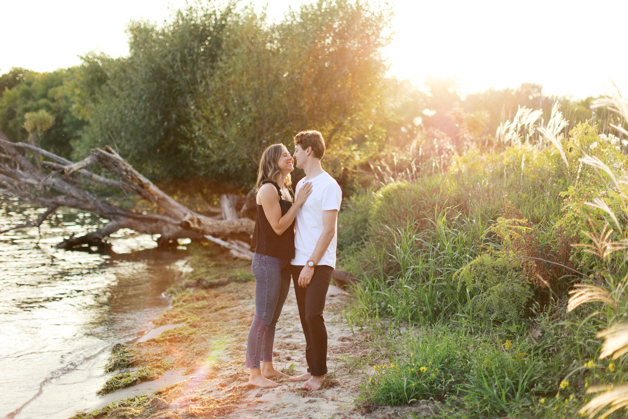 A couple stands close and smiles barefoot on the beach by Lake Minnetonka, the sunset casting a golden glow around them. Captured by Toly Dzyuba Photography.