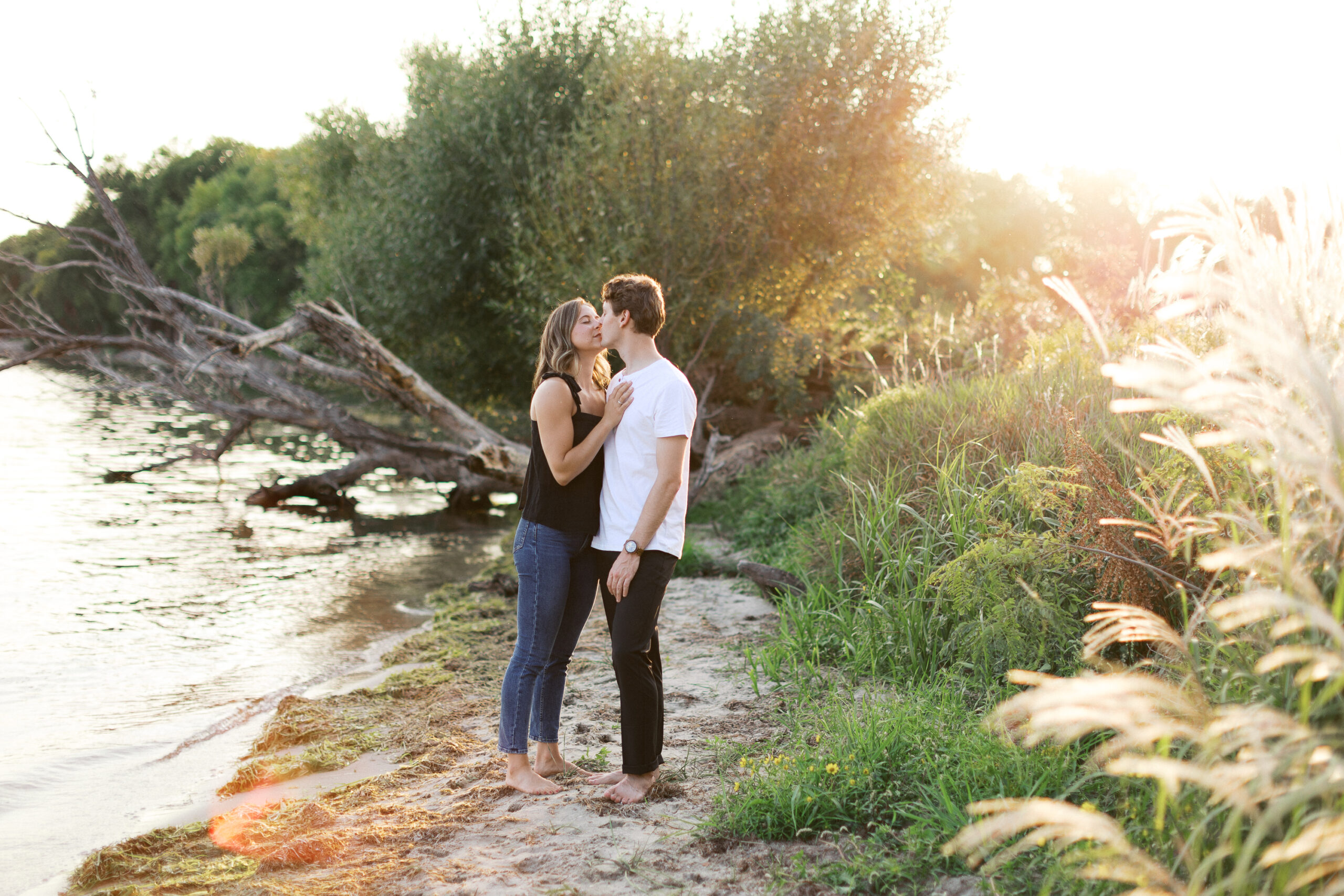 A romantic kiss as the couple stands barefoot on the beach by Lake Minnetonka, the sunset casting a golden glow around them. Captured by Toly Dzyuba Photography.