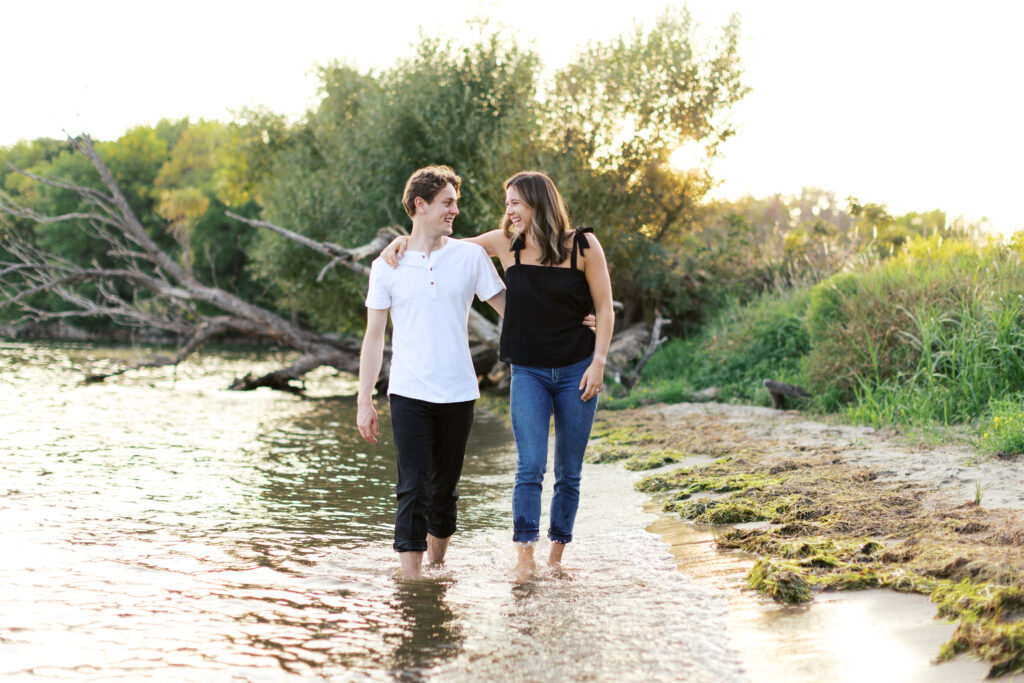 A couple walks on the beach as the sun sets over Lake Minnetonka. The golden light of the setting sun bathes the two in its warm glow. They are walking hand-in-hand in the shallow water. Photo by Toly Dzyuba Photography.