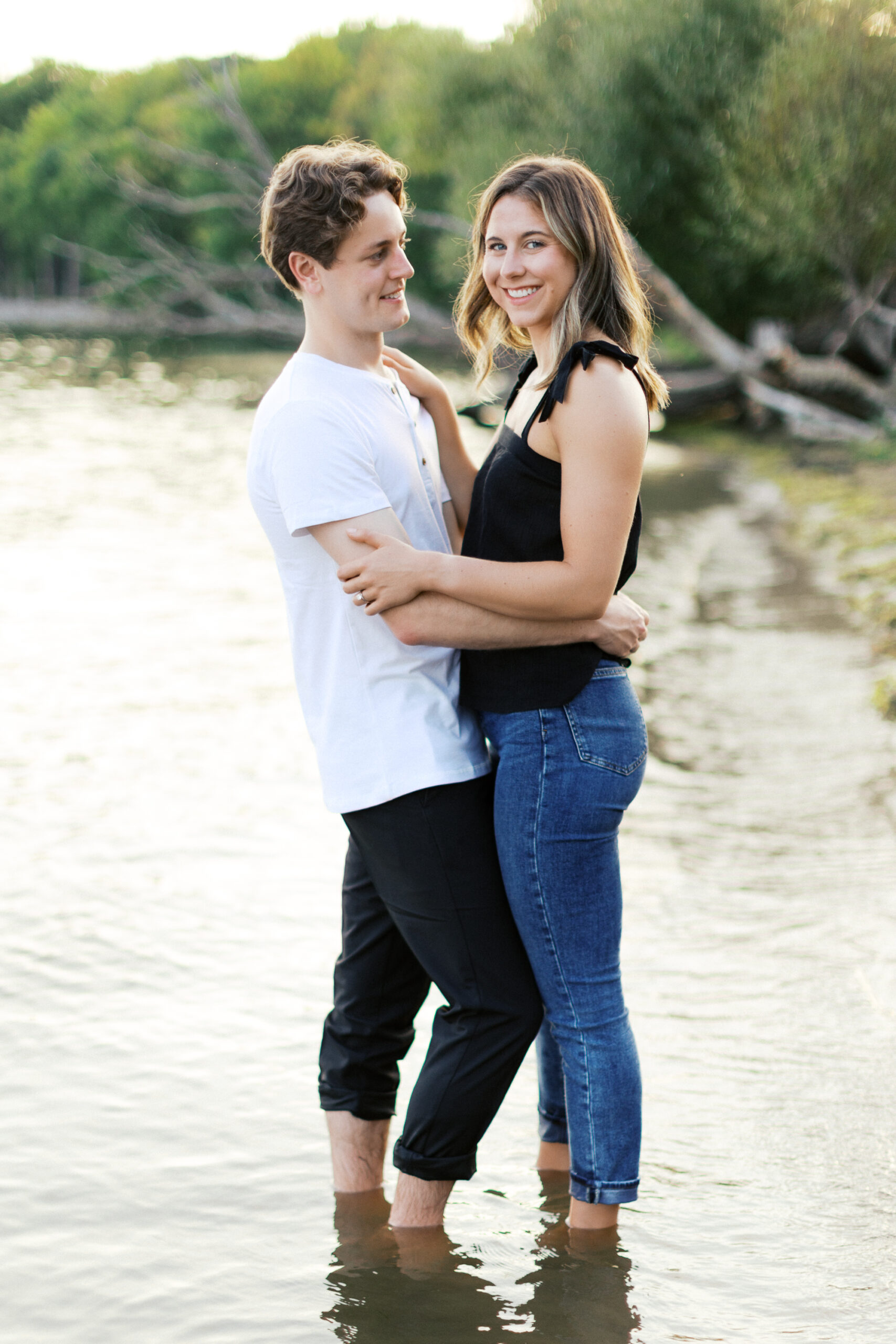 A romantic sunset moment at Noerenberg Gardens as the couple stands in the lake, their feet in the water. He gazes at her while she smiles softly at the camera, surrounded by golden light. Captured by Toly Dzyuba Photography.