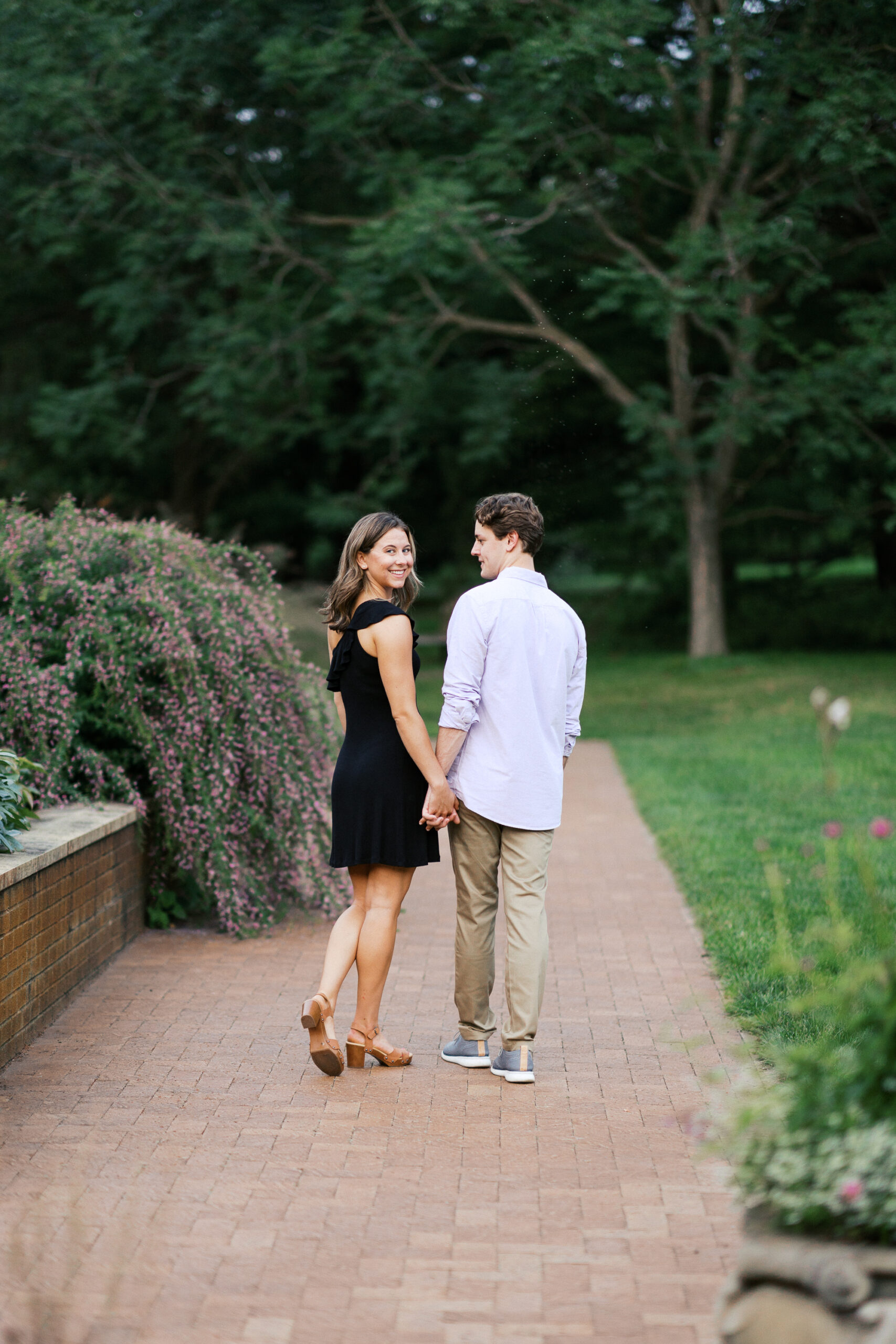 Couple is walking in the park, she is wearing a black dress. He is looking at her while she looks at the camera with a smile.