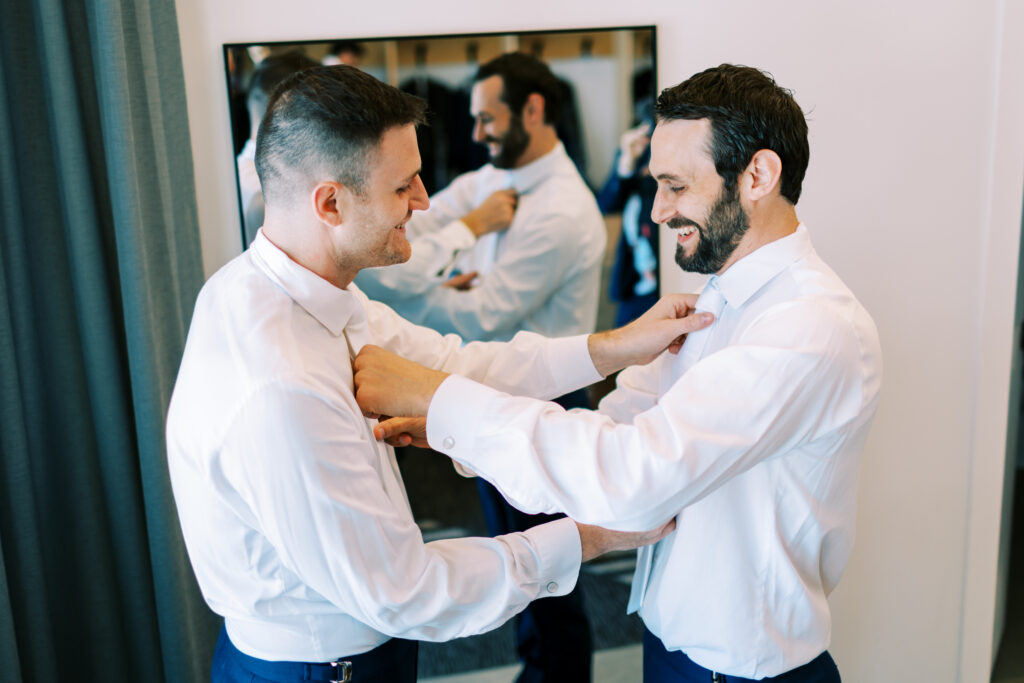 The brother of the groom is standing next to him, and they are about to fasten their dress shirts together.