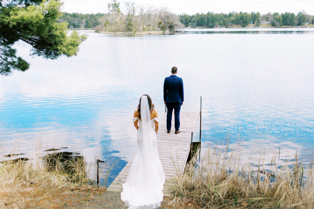 The bride approaches the groom from behind, standing on the lake dock with water on both sides of the dock. She and the groom are getting ready to see each other for the first time for the first look of the day.