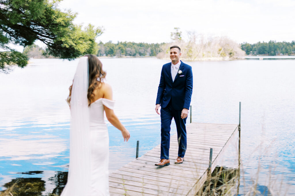 This picture shows a groom's reaction when he sees his bride for the first time. He is wearing a tuxedo and appears to be standing on a dock, and he is very excited to see her.