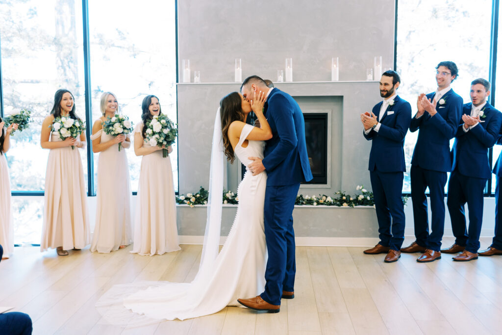 Bride and groom kiss at their wedding ceremony. The wedding party is cheering.