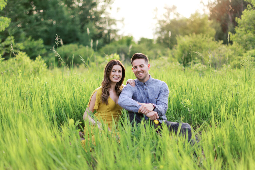 A couple sits in tall grass at Elm Creek Park Reserve in Maple Grove, both smiling as she rests her hand on his shoulder, captured by Minnesota wedding photographer Toly Dzyuba.