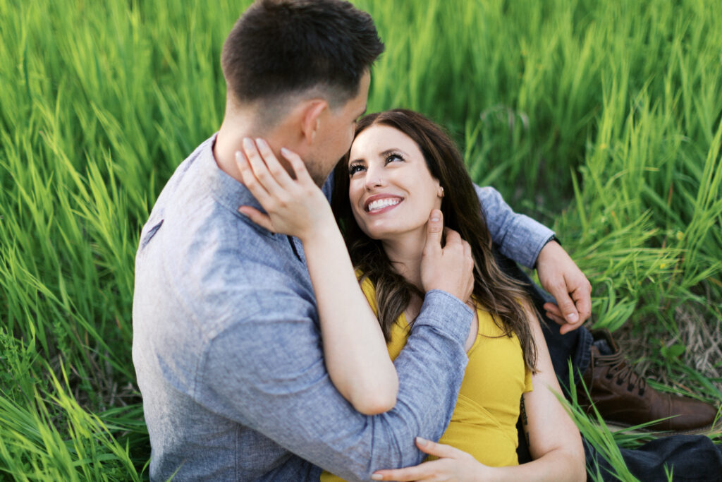 Woman lies on her partner’s lap in tall grass as he gazes into her eyes; she smiles and gently touches his hair.
