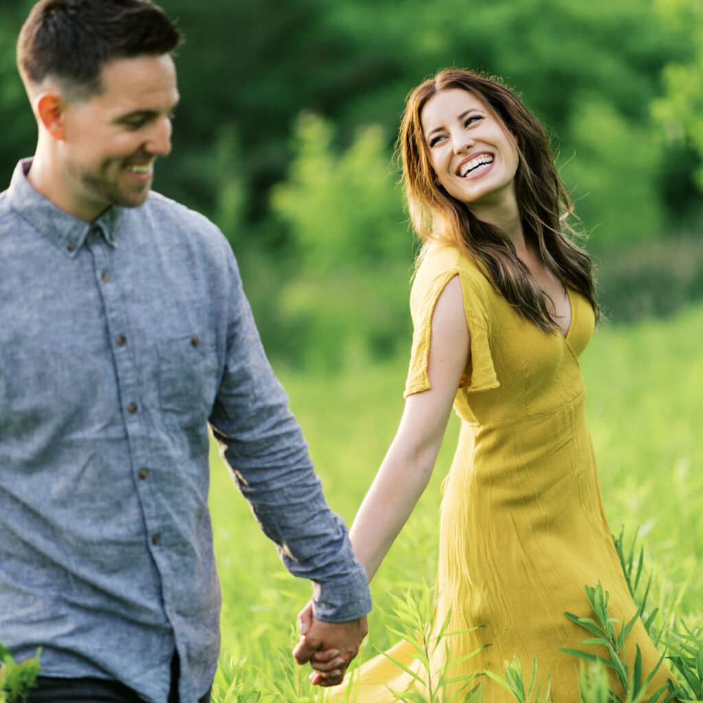 A couple strolls hand-in-hand through tall grass in Elm Creek Park, smiling joyfully as they share a carefree moment in nature.