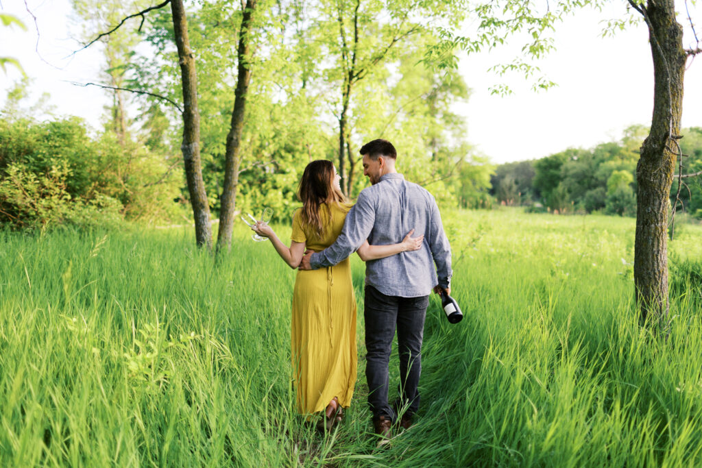 A couple walks through a grassy field with their backs turned, smiling as he hugs her waist and they hold a champagne bottle and glass under a sunset sky.