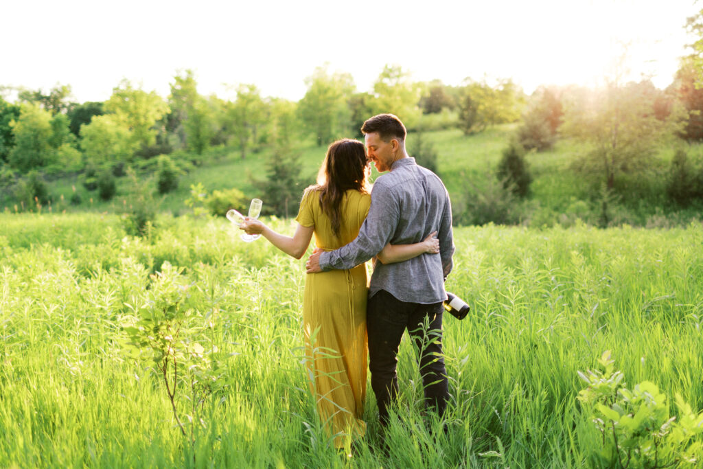 Couple walks through a sunlit field, holding champagne and a glass; he hugs her waist as they lean in for a sunset kiss.