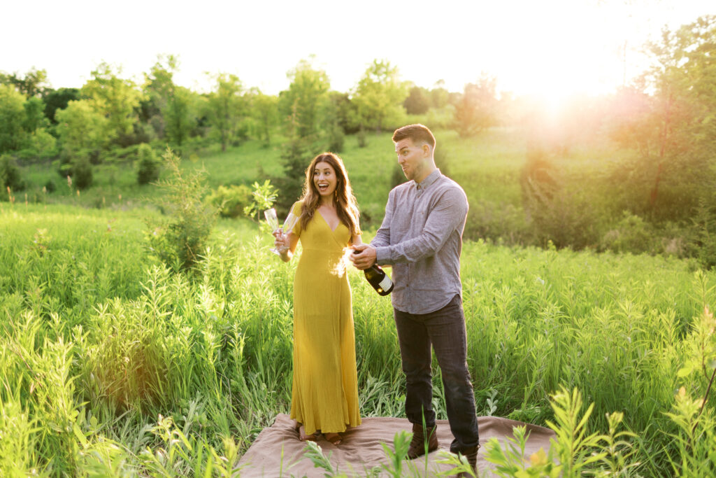 Couple in tall grass pop a champagne bottle, laughter and bubbly spray around them as she wears a yellow dress at Elm Creek Park Reserve.