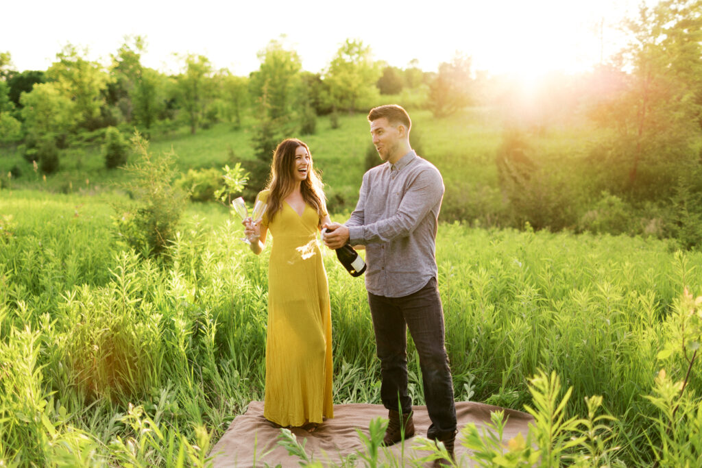 Couple opens the bottle of champagne in the glowing sunset of Elm Creek Park in Maple Grove. Surrounded by nature and grassy field. In the background, tall grass seems to whisper a message. It’s a romantic scene. They both in the joyful moment.