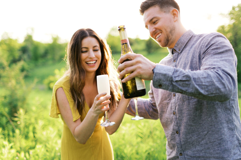 Couple in tall grass pop a champagne bottle, laughter and bubbly spray around them as she wears a yellow dress at Elm Creek Park Reserve.