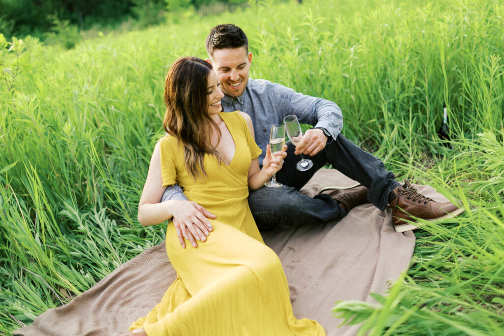 Couple sits on a blanket in tall grass, toasting with champagne glasses and smiling joyfully during a picnic in nature.