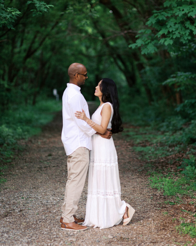 The couple stands together in white, looking into each other's eyes with smiles, surrounded by the serene beauty of a green tree tunnel.