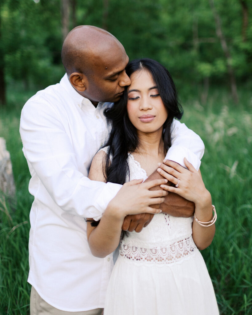 A gentleman kisses his girl on the head while hugging her from behind, capturing a tender moment of love and intimacy.