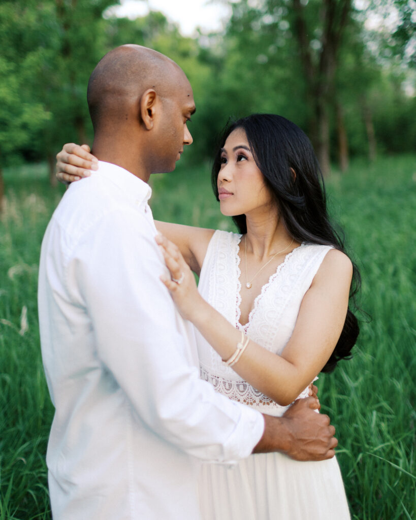 She wraps her arms around his shoulders as they look deeply into each other's eyes, surrounded by the beauty of nature at Elm Creek Park in Maple Grove.