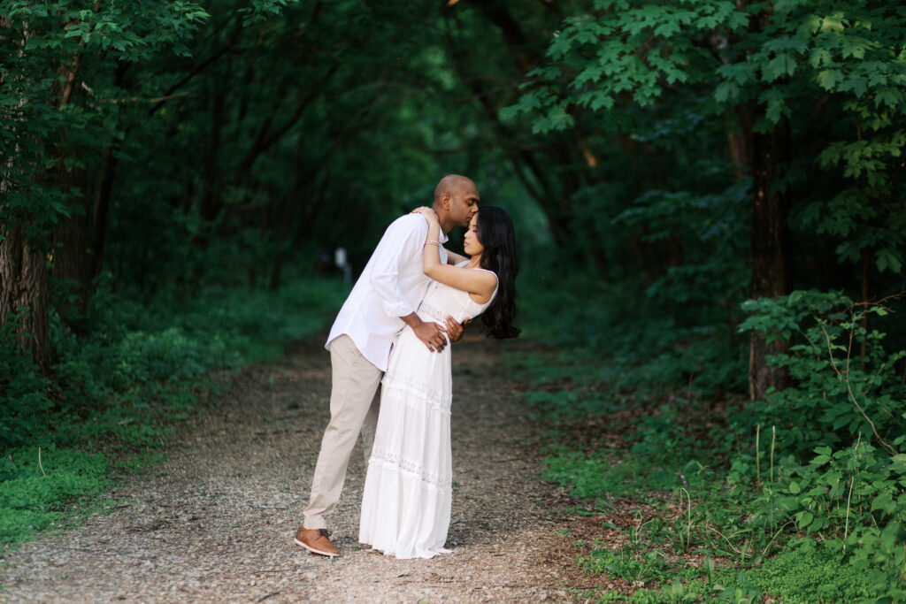 A man dips his woman and kisses her under a canopy of lush green trees, creating a romantic moment in a natural tunnel of foliage.