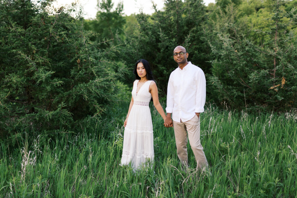 Couple holding hands and looking at the camera, standing together in a picturesque natural setting.
