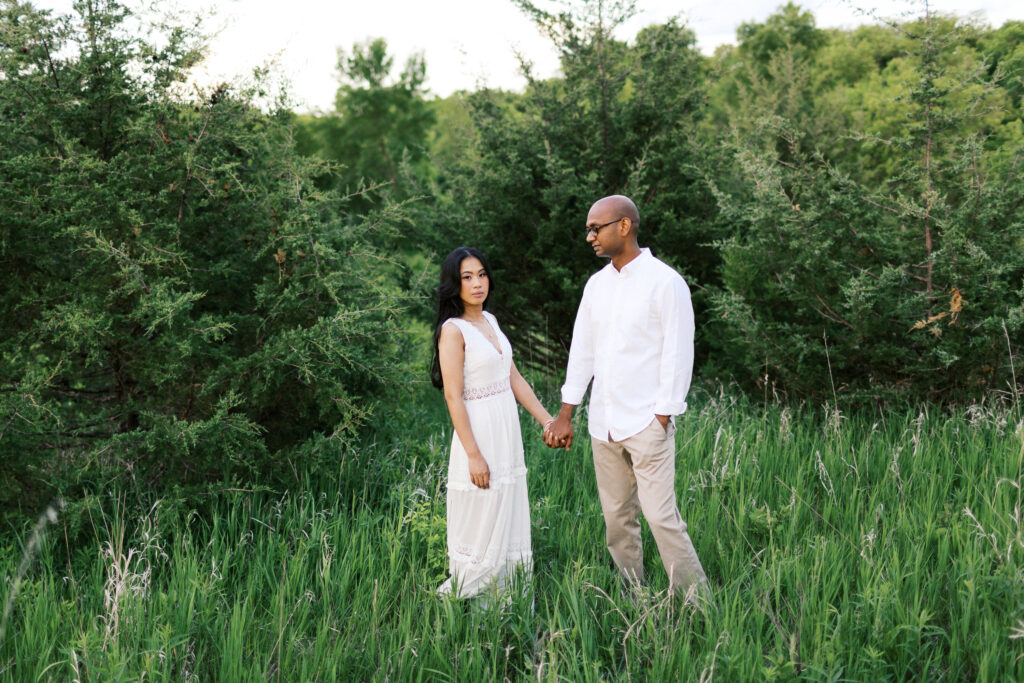 He holds her hand as she looks at the camera, surrounded by nature, capturing a beautiful and romantic moment.