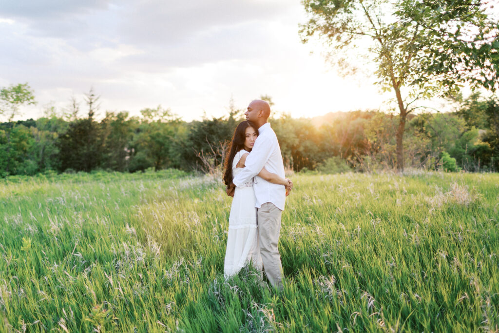 A man and woman stand in the tall grass, embracing as he gently kisses her hair in the golden sunset at Elm Creek Park, captured by Minnesota wedding photographer Toly Dzyuba.