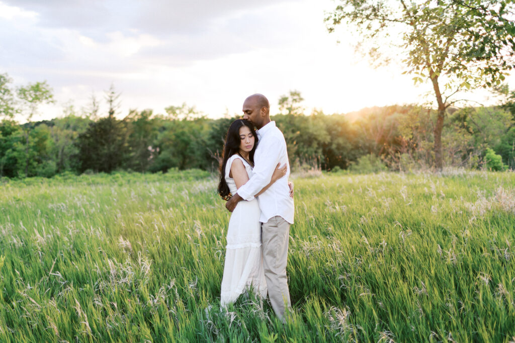 A man and woman stand in the tall grass, embracing as he gently kisses her hair in the golden sunset at Elm Creek Park, captured by Minnesota wedding photographer Toly Dzyuba.