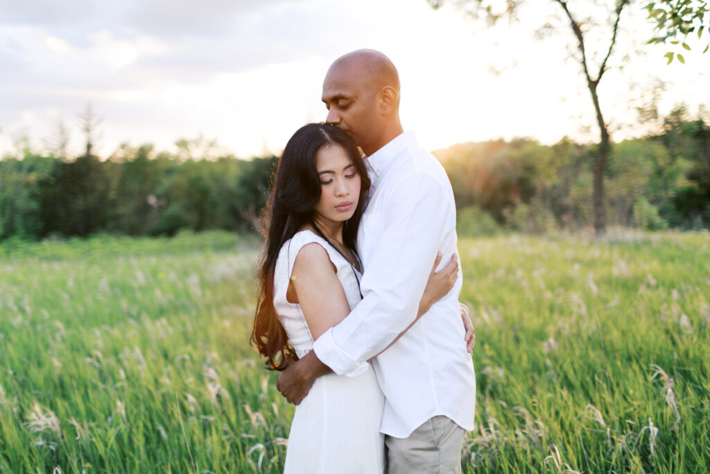 A man and woman stand in the tall grass, embracing as he gently kisses her hair in the golden sunset at Elm Creek Park, captured by Minnesota wedding photographer Toly Dzyuba.