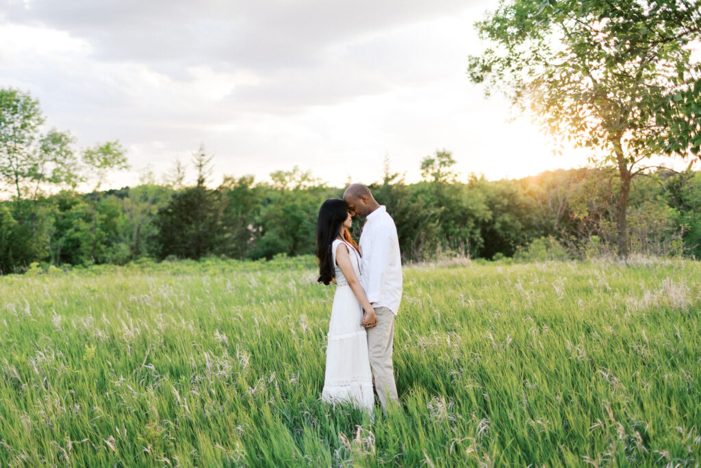 They hold hands and rest their heads together in the golden sunset, surrounded by tall grass, sharing a close and candid moment of love.