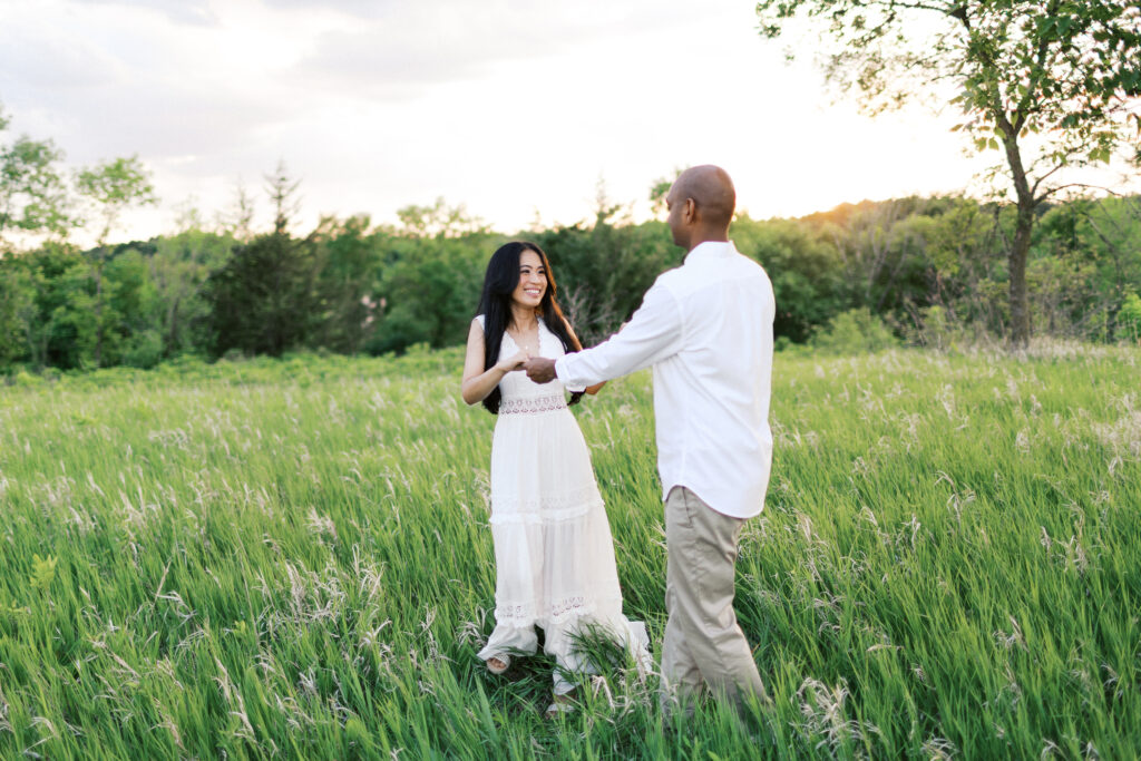 They laugh and dance in the tall grass at sunset, spinning in a circle while holding hands, filled with pure joy and love.