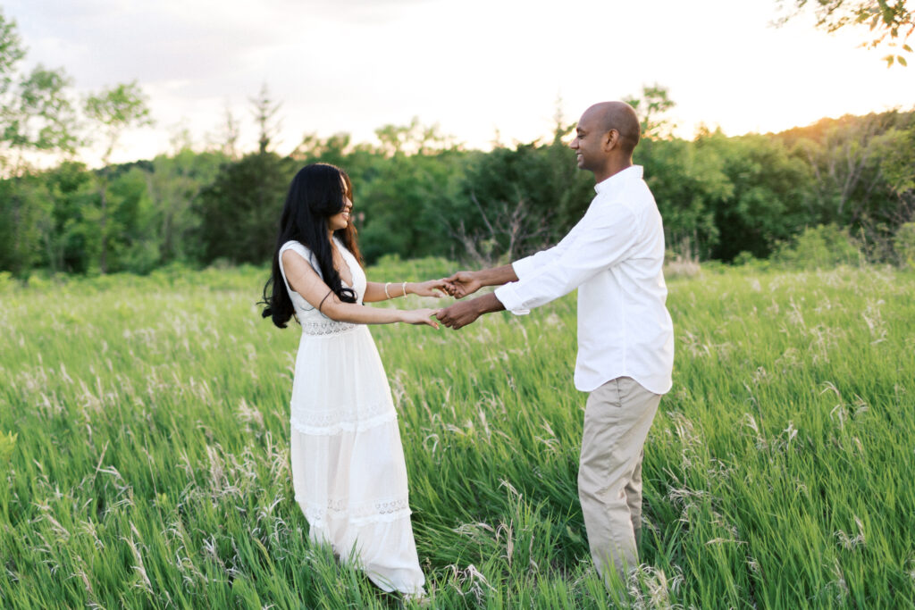 They laugh and dance in the tall grass at sunset, spinning in a circle while holding hands, filled with pure joy and love.